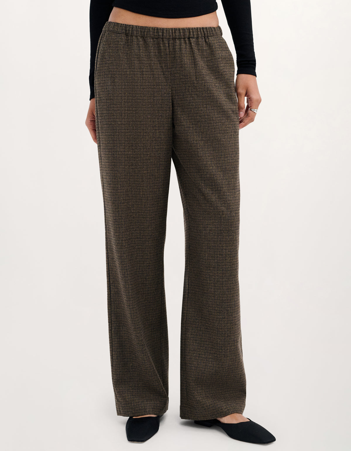 A person wearing loose, wide-leg trousers in a dark brown and olive-toned textured pattern, paired with a black long-sleeve top and black flats, photographed against a plain light background.