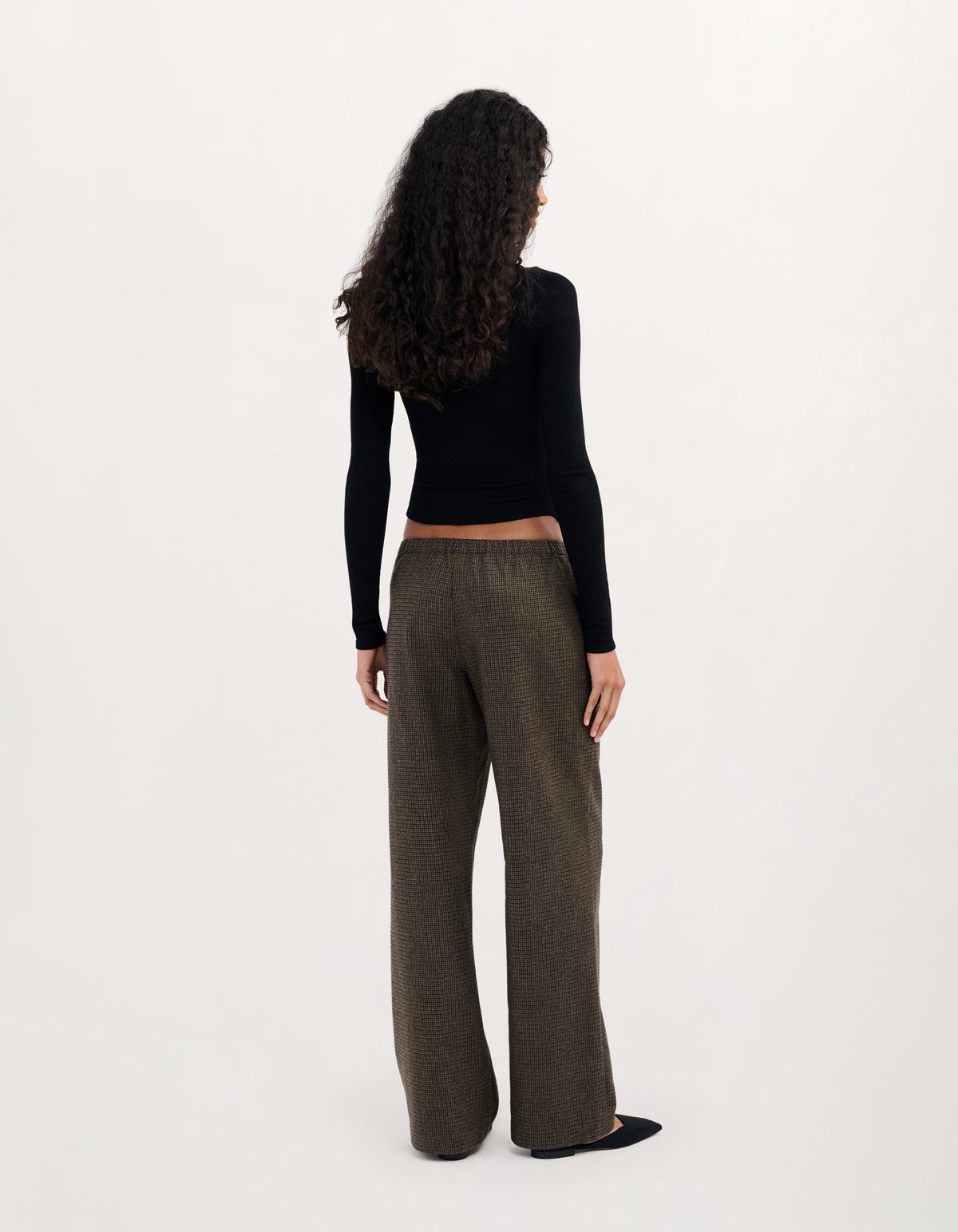 A model shown from the back wearing a black long-sleeve crop top and dark brown wide-leg trousers against a plain white studio background.