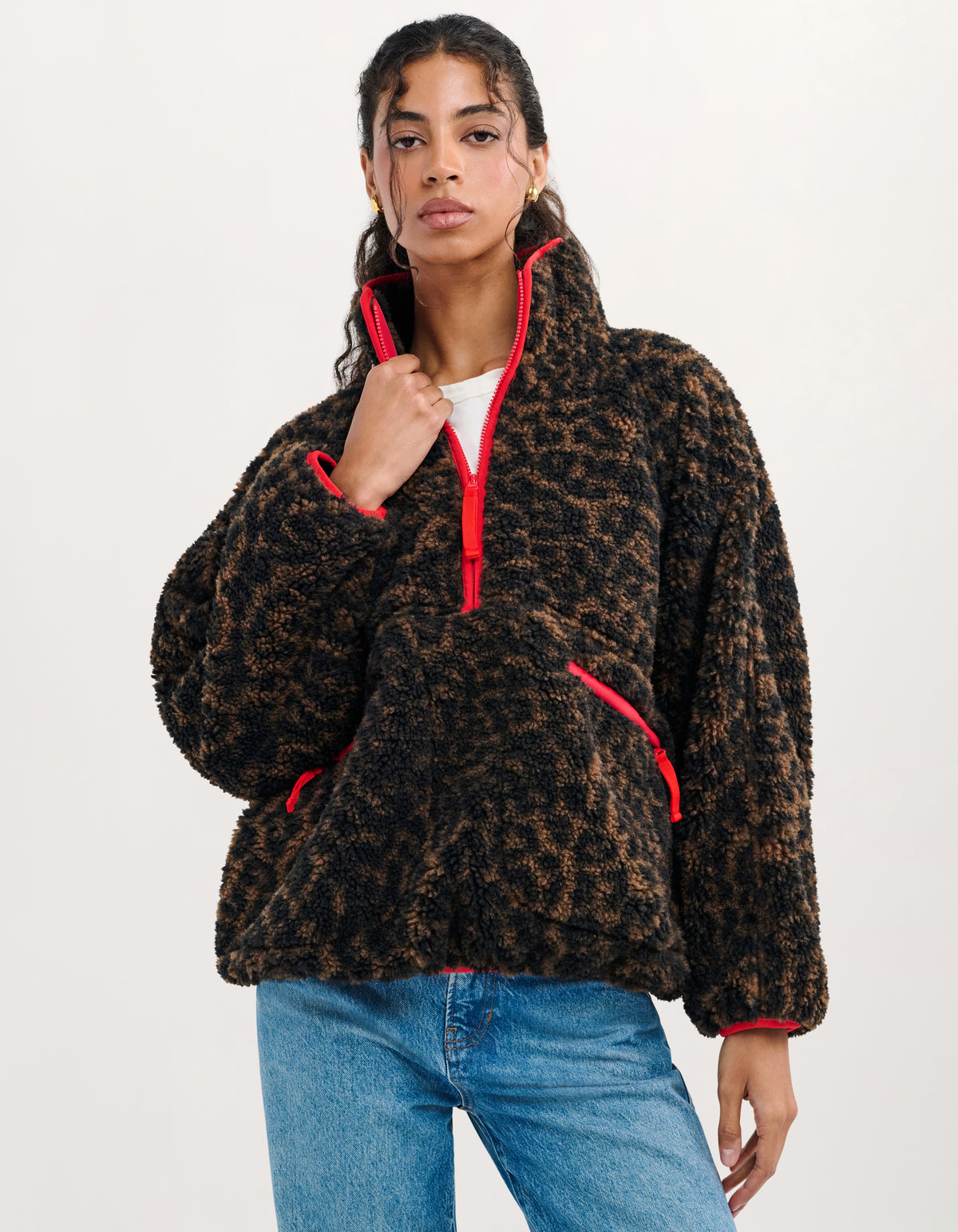A woman wearing a brown and black leopard-print fleece pullover with bright red trim and zipper details, paired with blue jeans, against a plain white studio background.
