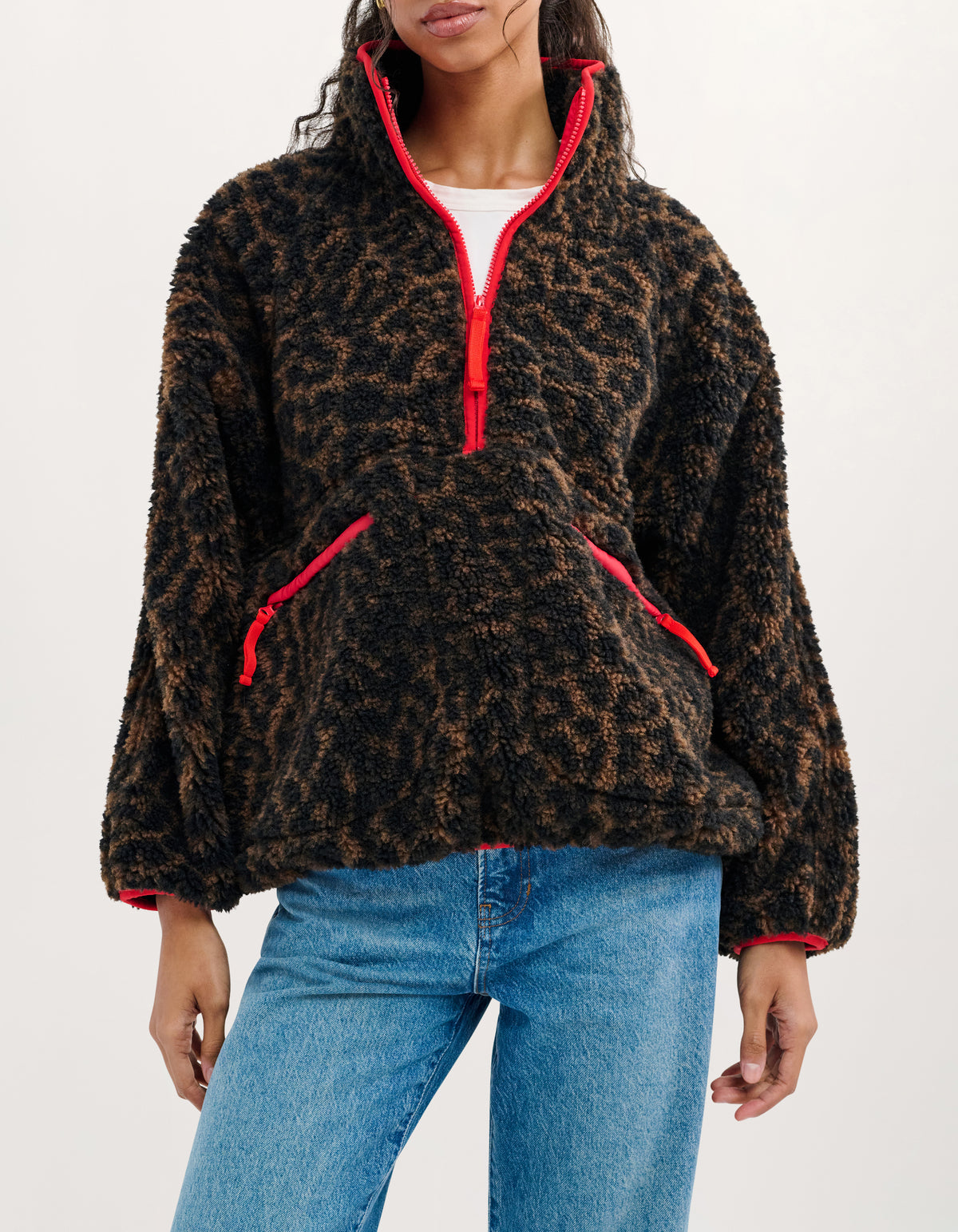 A woman wearing a brown and black leopard-print fuzzy quarter-zip fleece pullover with bright red trim and zippers, paired with blue jeans, against a plain white studio background.