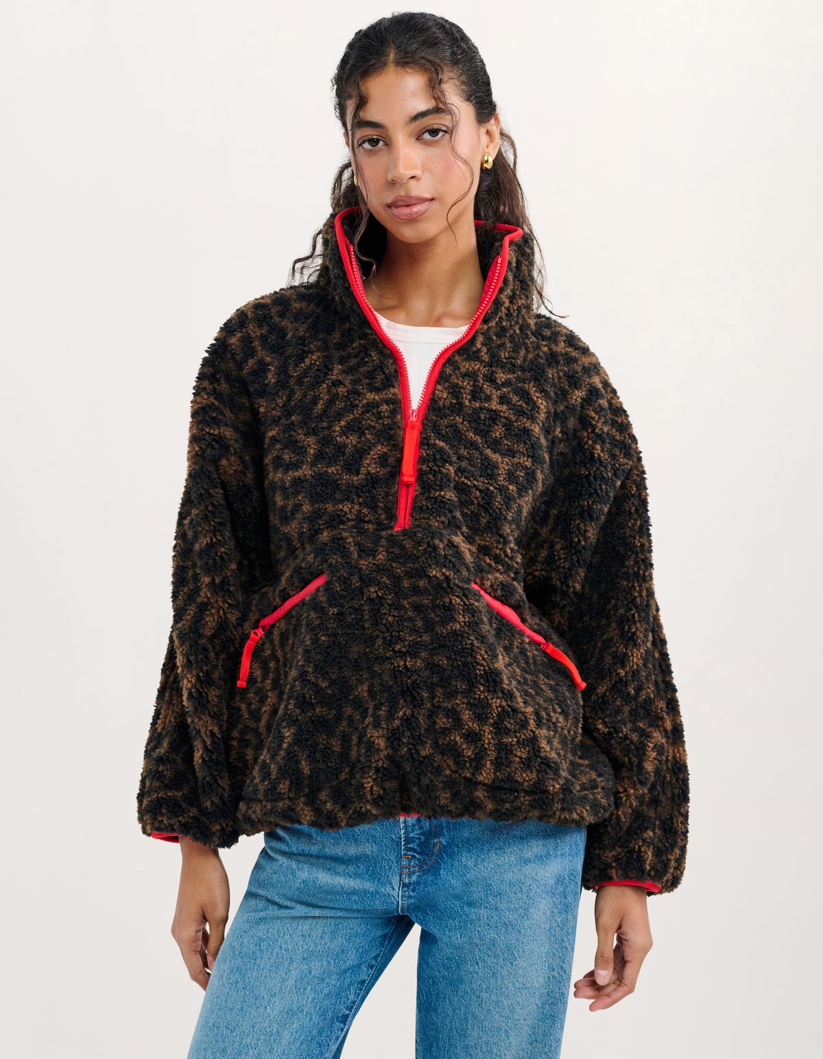 A woman modeling a black and brown leopard-print fleece pullover with bright red trim and a red zipper, paired with blue jeans against a plain white studio background.