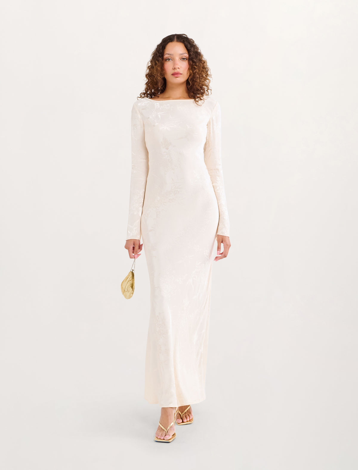 A woman modeling a long-sleeved, floor-length cream lace dress with a boat neckline, paired with a small gold clutch and gold strappy heels, against a solid off-white background.