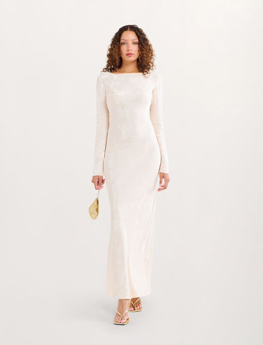 A woman modeling a long-sleeved, floor-length cream lace dress with a boat neckline, paired with a small gold clutch and gold strappy heels, against a solid off-white background.