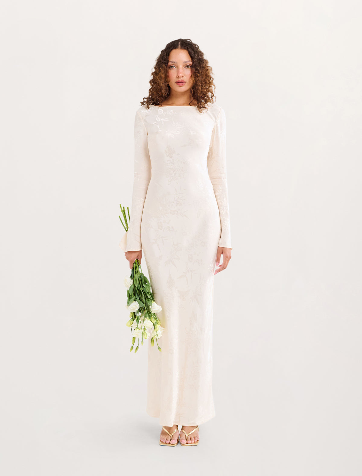 A woman stands against a plain light background wearing a long-sleeved cream maxi dress with a boat neckline and subtle tonal floral embroidery. She holds a small bouquet of white flowers and wears metallic sandals.