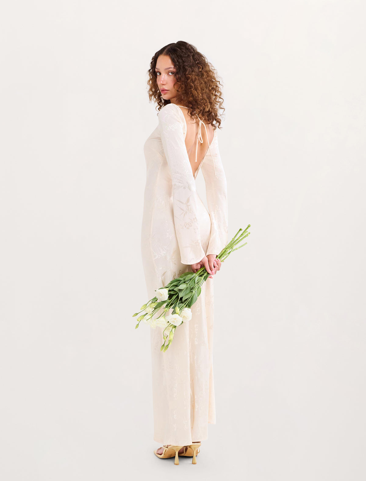 A woman with curly brown hair stands in profile wearing a long-sleeved, off-white lace dress with a deep back and holding a bouquet of white flowers, set against a plain white studio background.