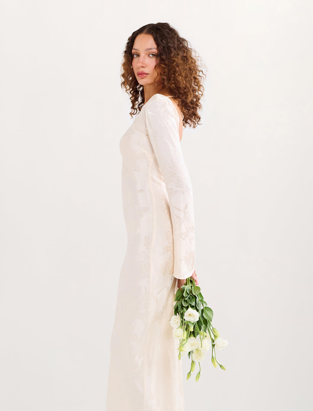 A woman with curly brown hair stands in profile wearing a long-sleeved cream lace dress and holding a small bouquet of white flowers against a plain white studio background.