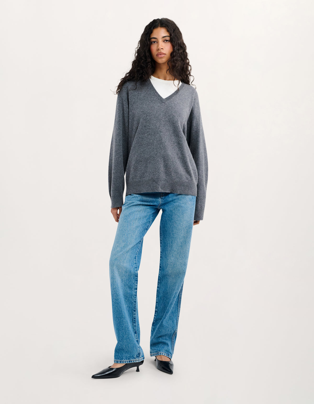 A model stands against a plain light background wearing a dark gray v-neck knit sweater over a white undershirt, blue jeans, and black pointed flats.