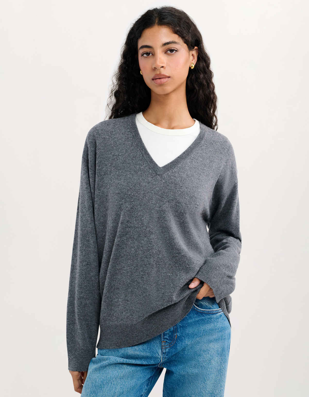 A woman modeling a heather gray long-sleeve V-neck sweater layered over a white crewneck shirt and paired with blue jeans, against a plain off-white studio background.