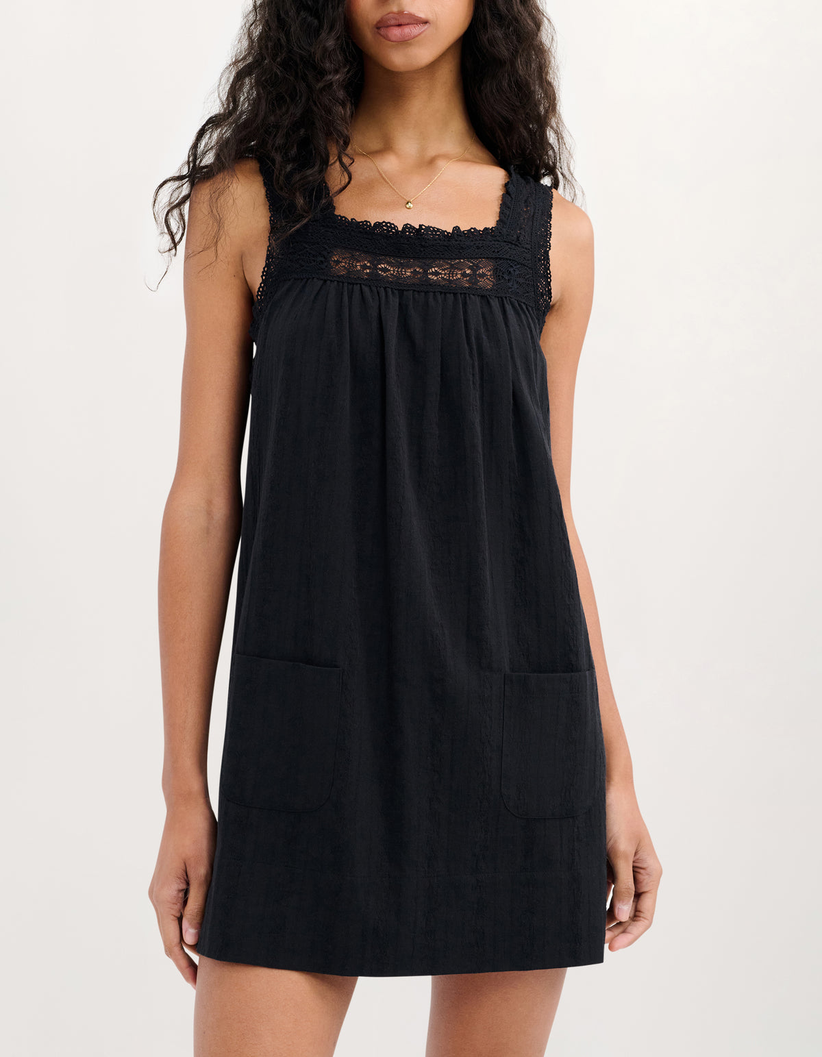 A woman wearing a black sleeveless linen dress with a lace-trimmed square neckline and two front patch pockets, photographed against a plain light background.