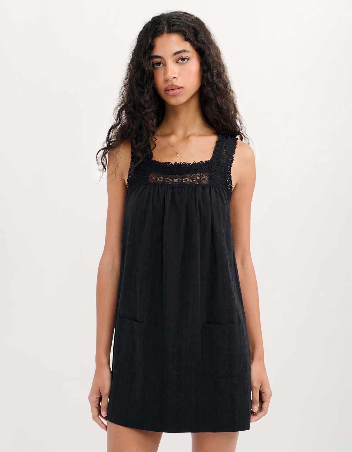 A woman modeling a black sleeveless mini dress with a square neckline, black lace trim across the chest, and two front patch pockets, set against a plain light gray studio background.