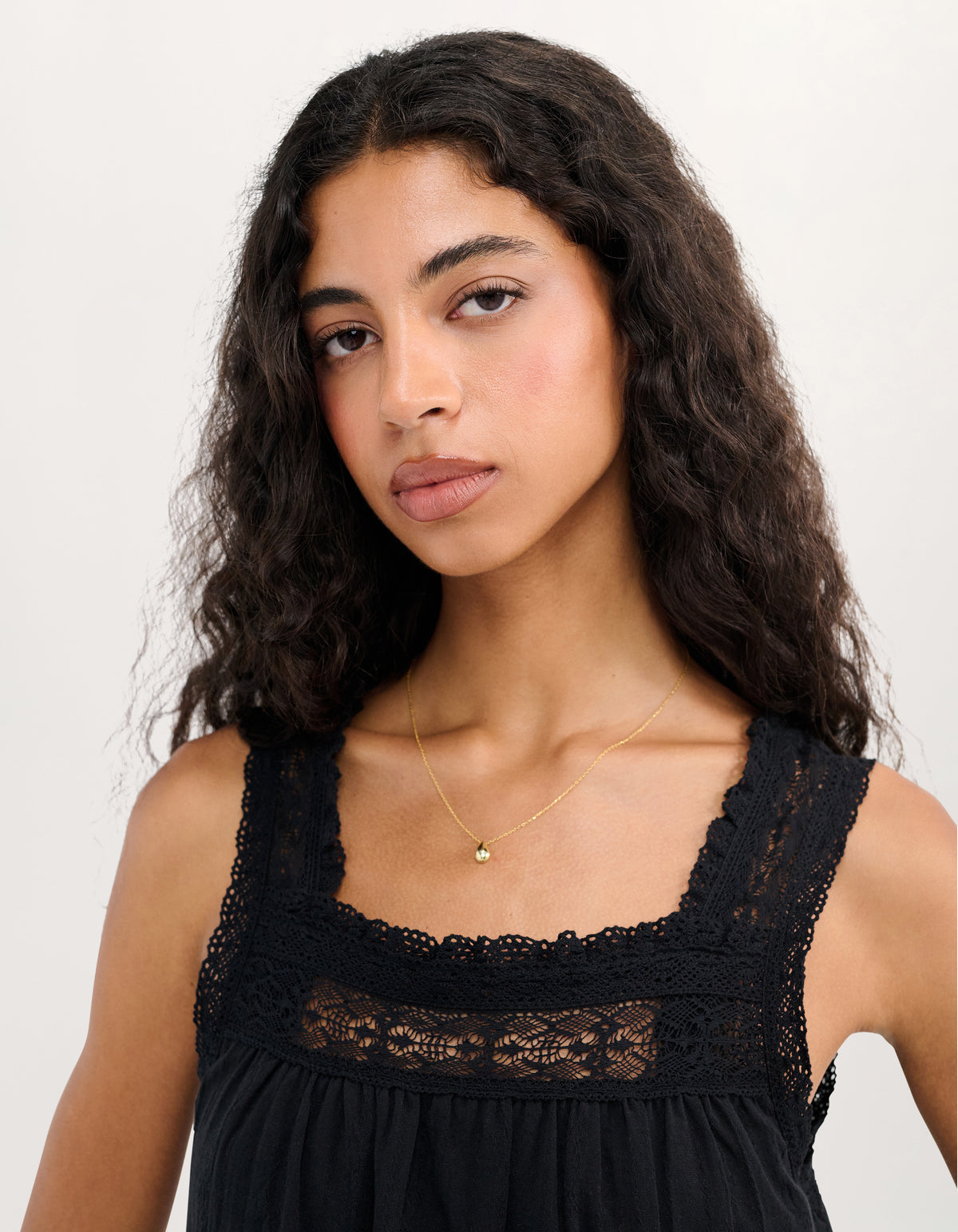 A woman with long, wavy dark hair is shown from the chest up, wearing a black lace-trimmed top and a delicate gold necklace with a small gold bead pendant, set against a plain light background.
