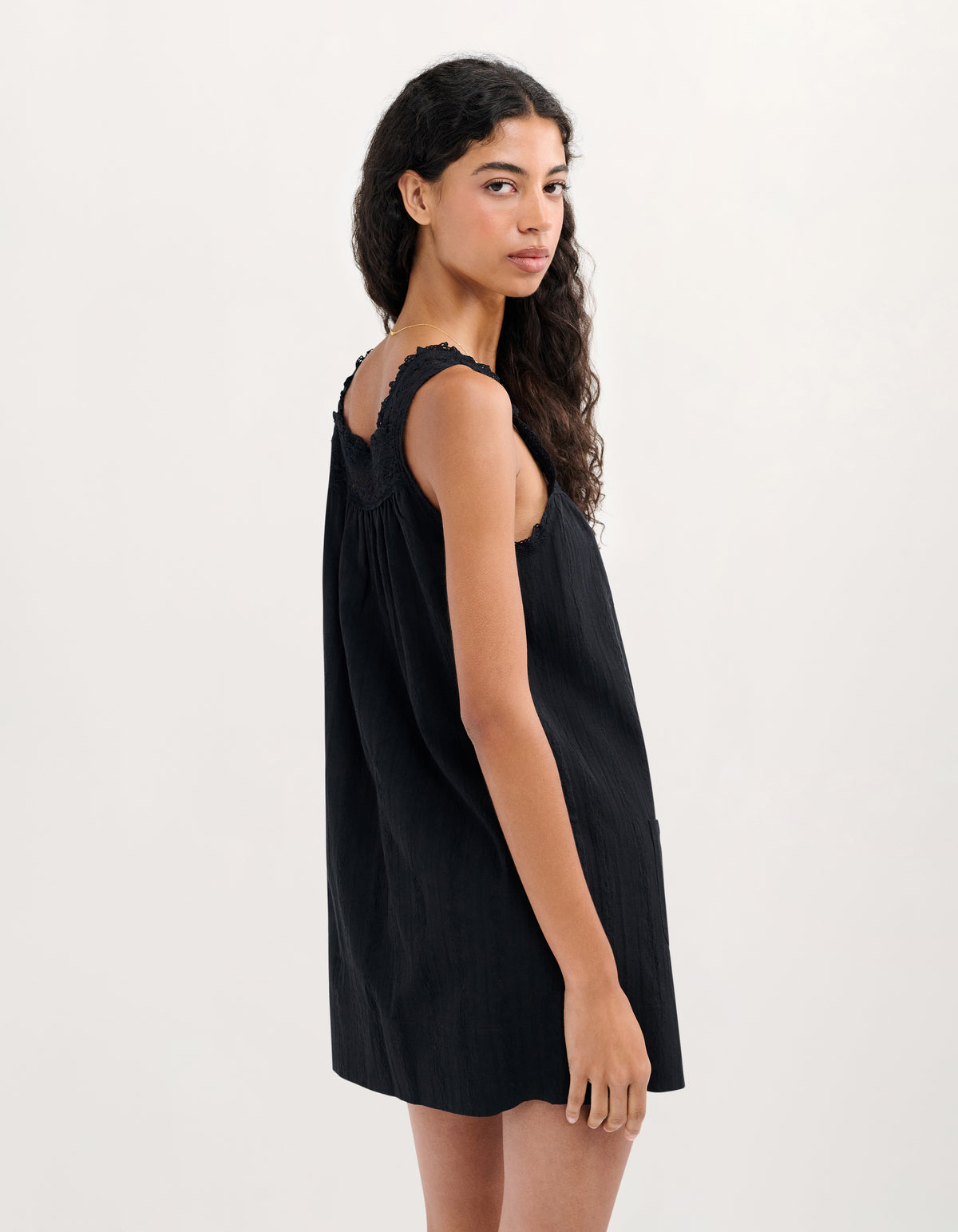A woman modeling a sleeveless black dress with a ruffled shoulder strap and gathered side pockets, photographed against a plain light gray studio background.