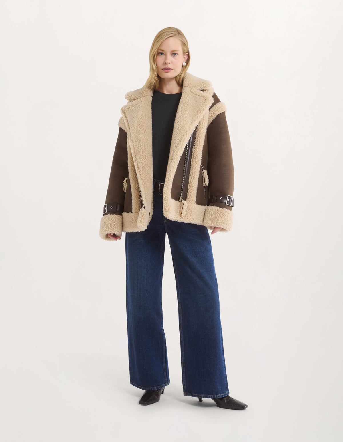 A model stands against a plain light gray studio background wearing a brown and beige shearling-style moto jacket over a black top and dark blue wide-leg jeans.