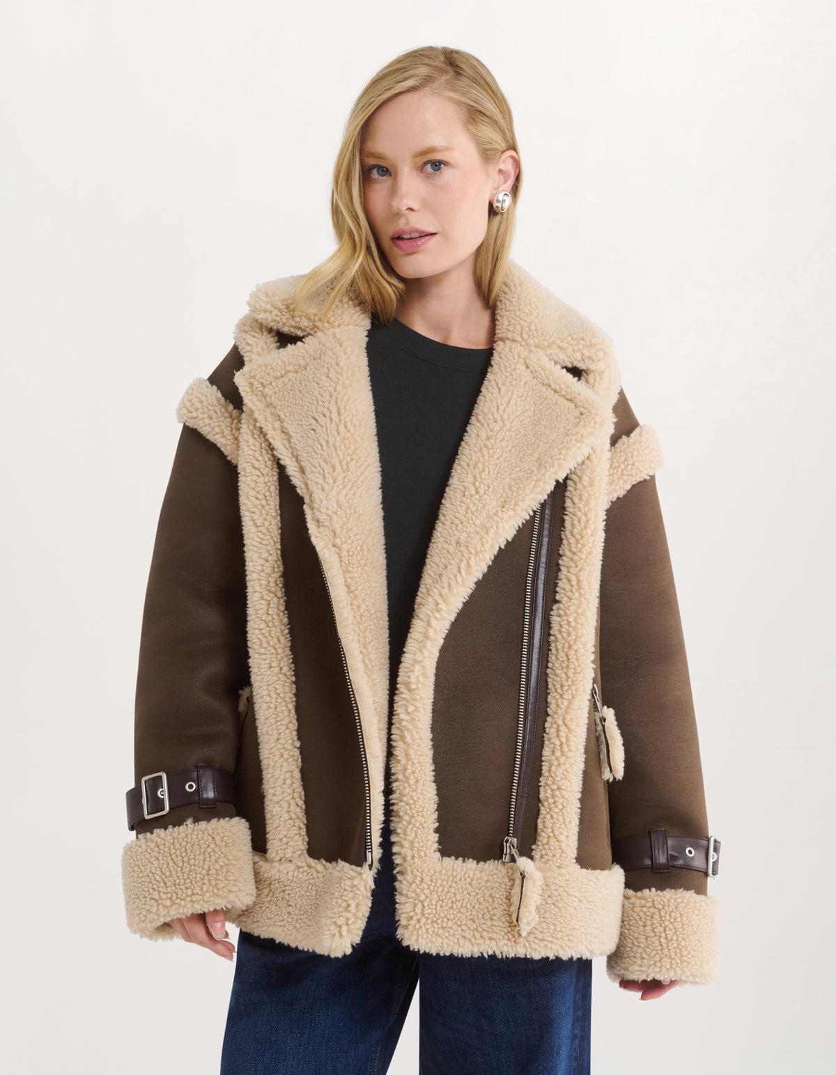 A woman wearing a brown and beige shearling-lined aviator jacket over a black top and blue jeans, photographed against a clean off-white studio background.