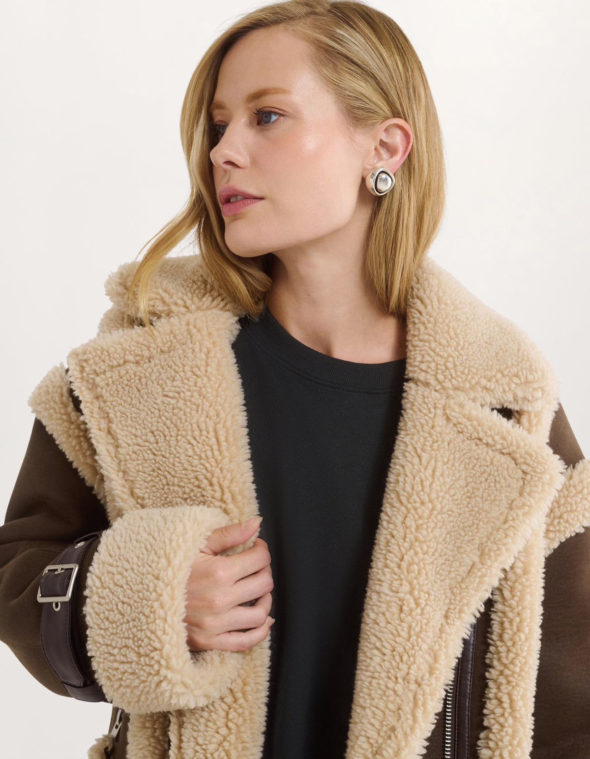 A woman wearing a beige sherpa fleece jacket with a brown leather-look sleeve and collar, layered over a black top. She is shown in a close-up profile against a clean white background.