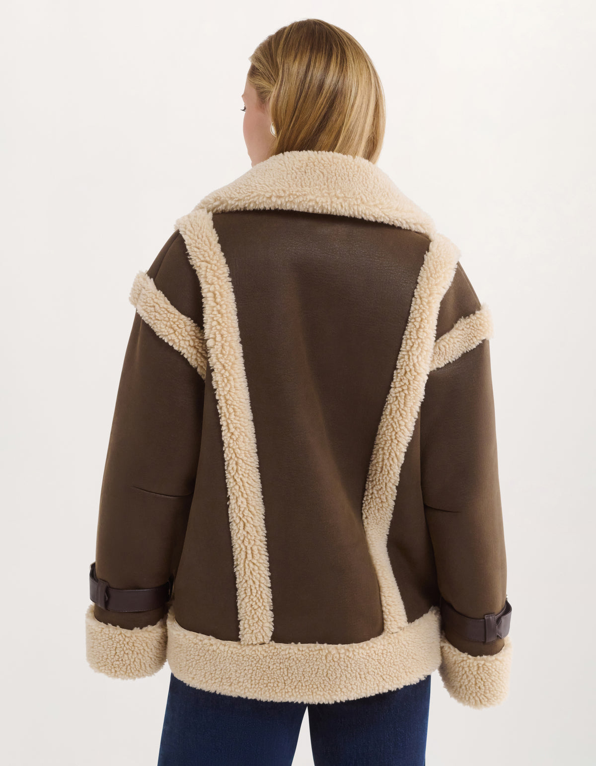 A woman viewed from the back wearing a brown shearling-lined jacket with cream-colored fleece trim and dark brown cuffs, paired with blue jeans against a plain white studio background.