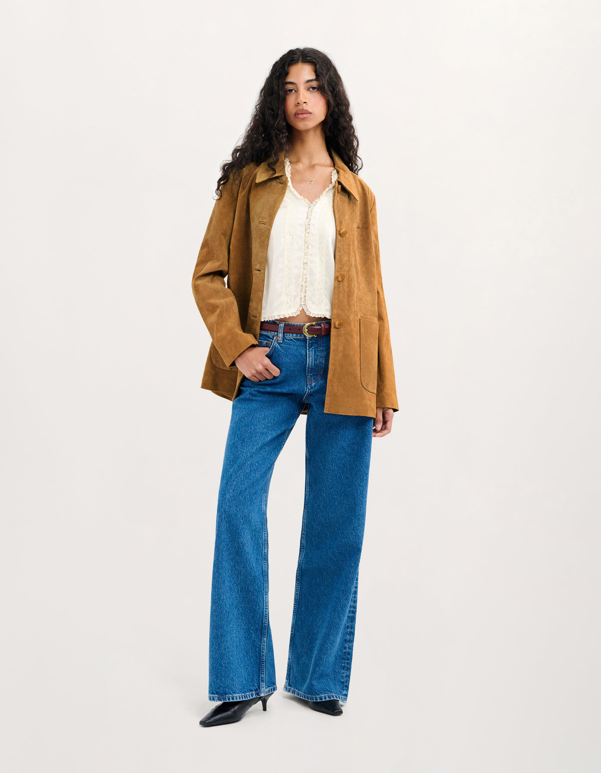 A model stands against a plain light background wearing a tan overshirt, a white lace-trim blouse, blue wide-leg jeans, and black pointed shoes.