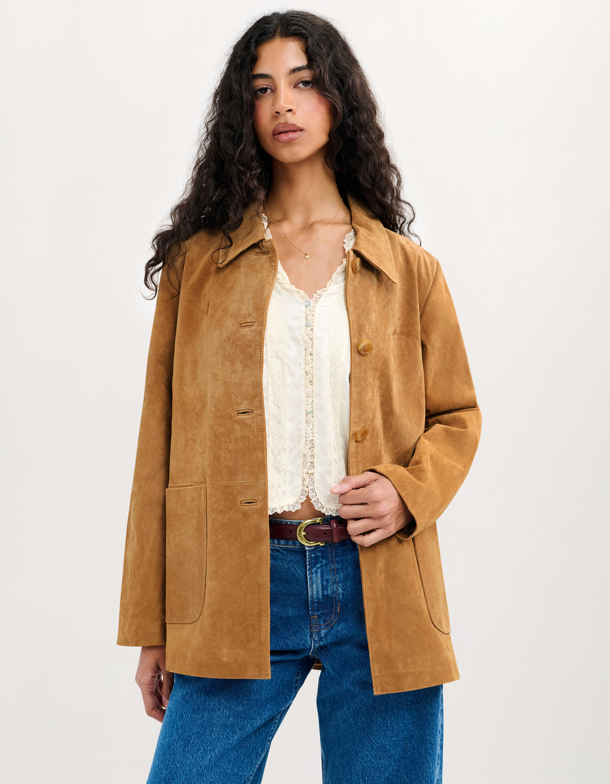 A woman modeling a tan suede jacket over a white lace-trim blouse and blue jeans, photographed against a plain light background.