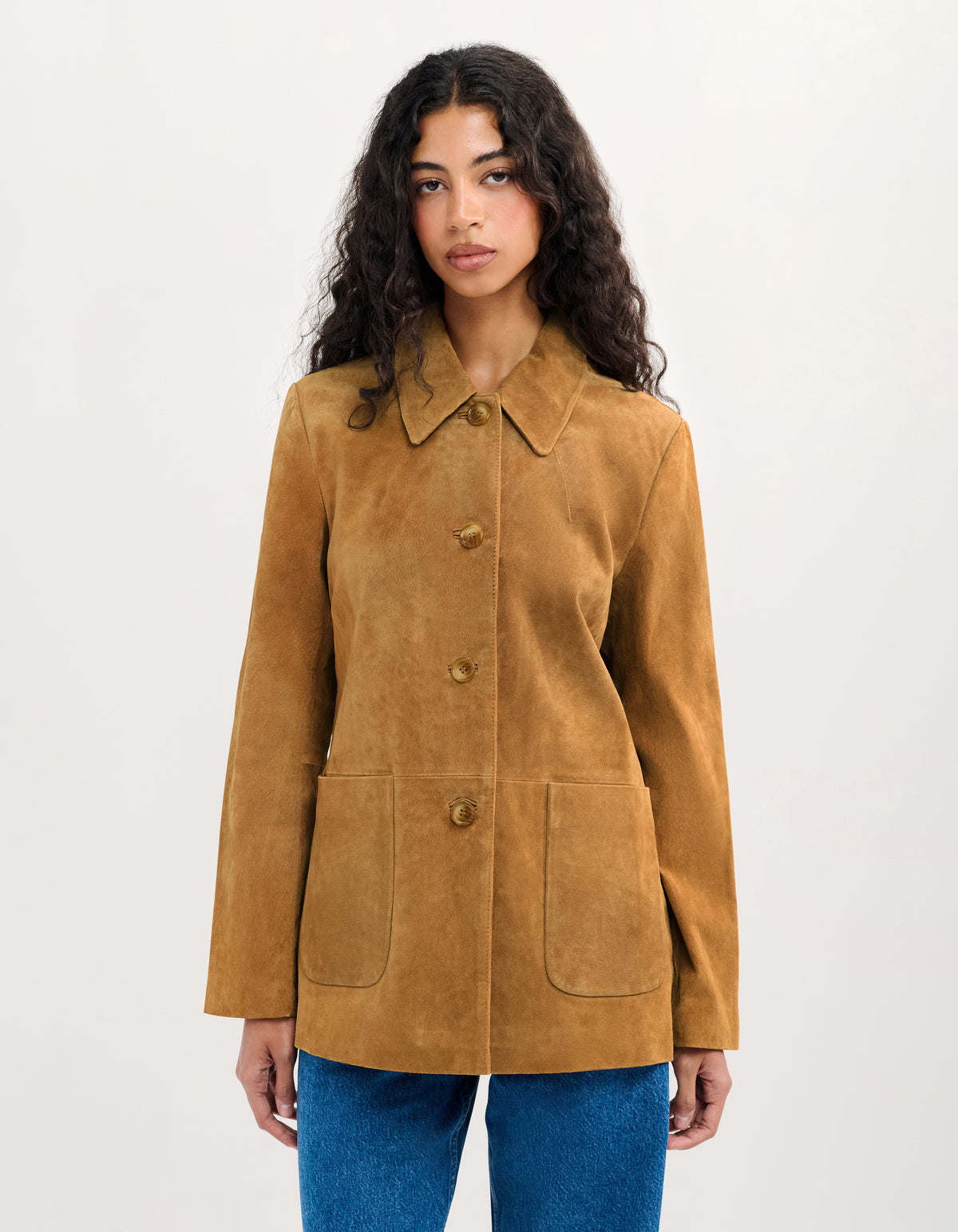 A woman modeling a tan suede jacket with a collar, three front buttons, and two large front patch pockets, paired with blue jeans against a plain light background.