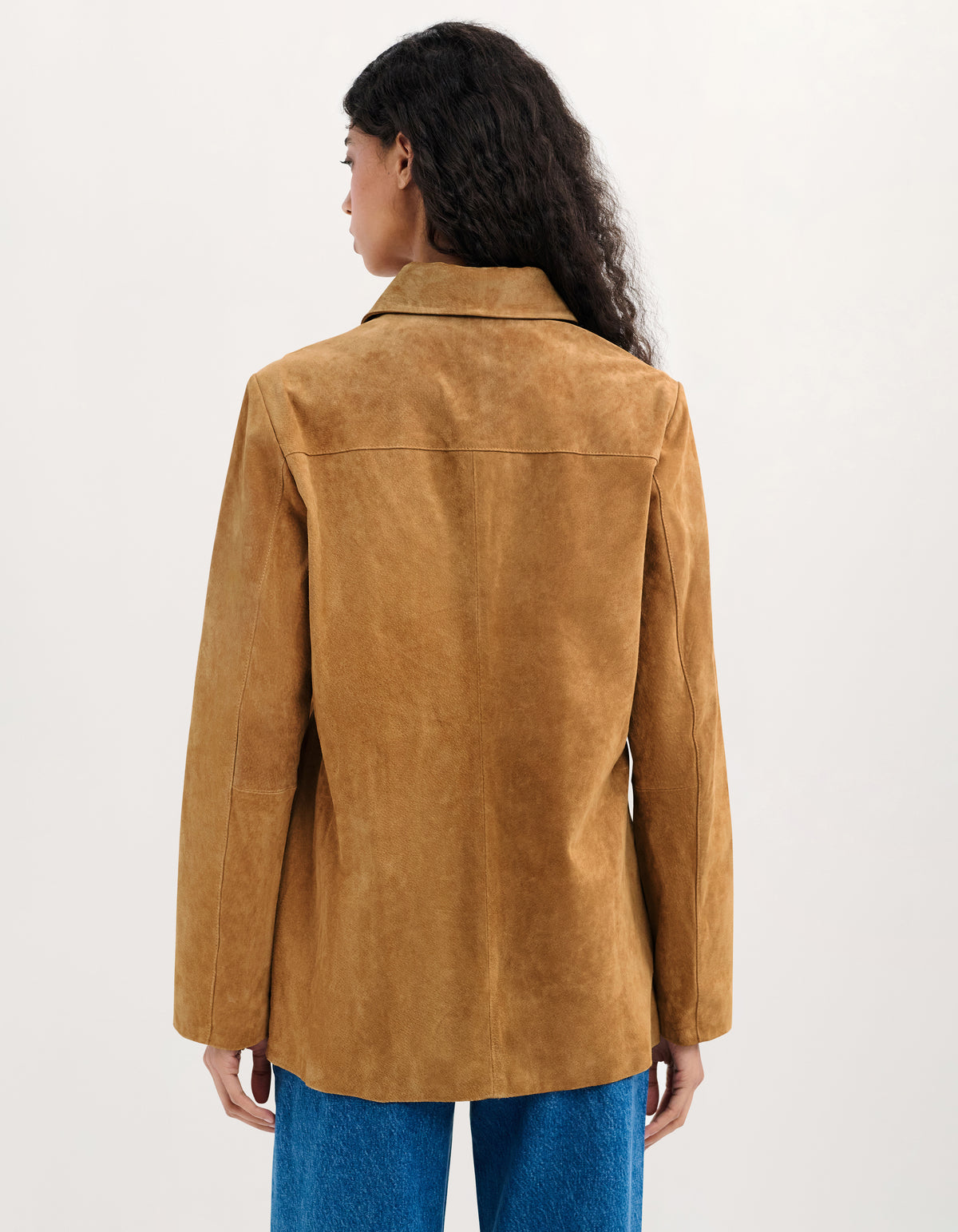 A woman shown from the back wearing a tan suede jacket and blue jeans against a plain white studio background.